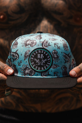 A tattooed person holds the Relentless Betrayal Sea Swallows the Weak PVC Snapback, a turquoise and black hat with tattoo-inspired illustrations and a bold round patch.