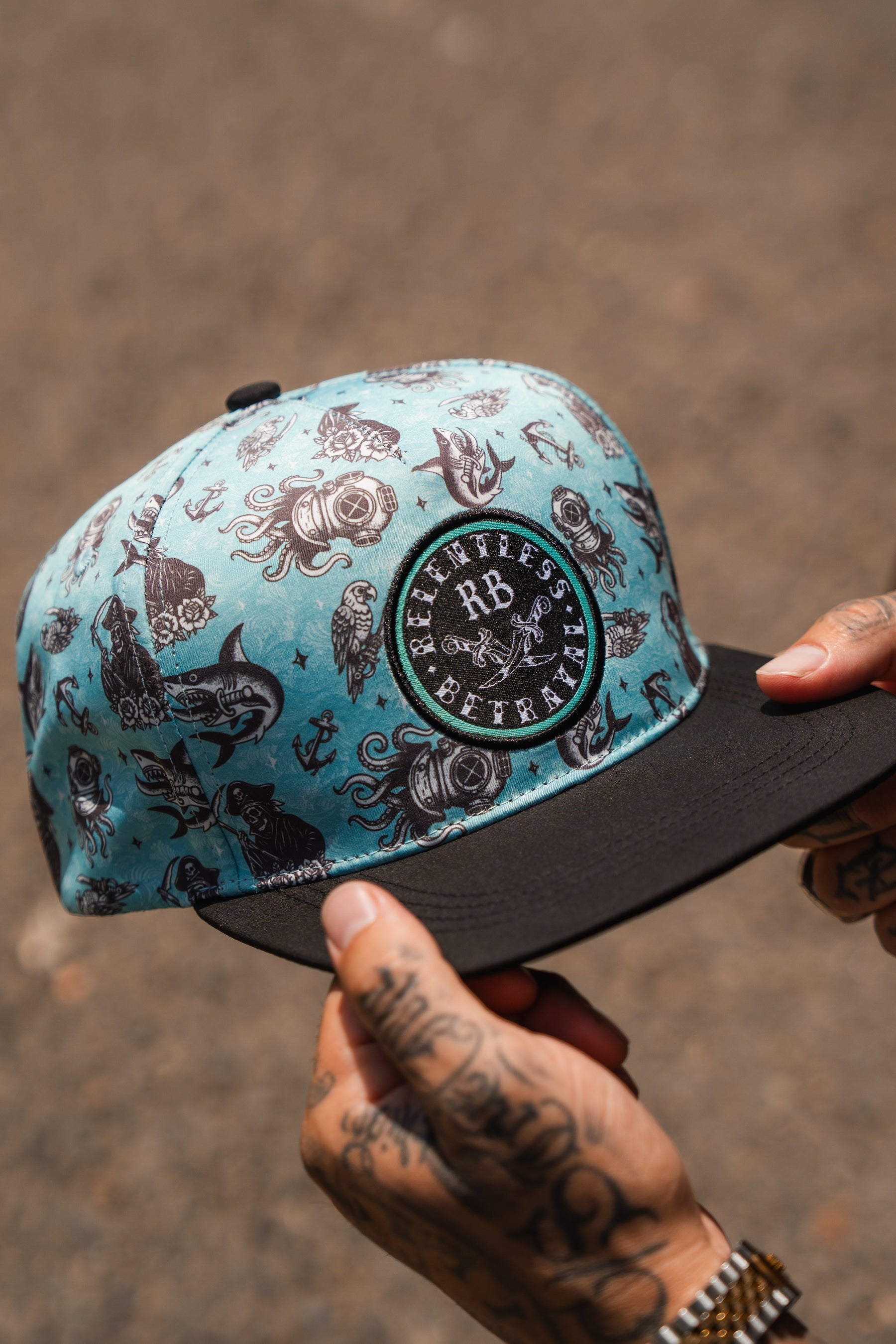 A tattooed person holds the Relentless Betrayal Sea Swallows the Weak PVC Snapback, a teal and black water-resistant hat with nautical designs and a front patch displaying "Relentless Betrayal"—ideal for those who embrace the pirate lifestyle.