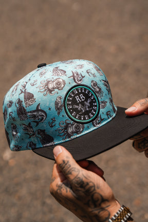 A tattooed person holds the Relentless Betrayal Sea Swallows the Weak PVC Snapback, a teal and black water-resistant hat with nautical designs and a front patch displaying "Relentless Betrayal"—ideal for those who embrace the pirate lifestyle.