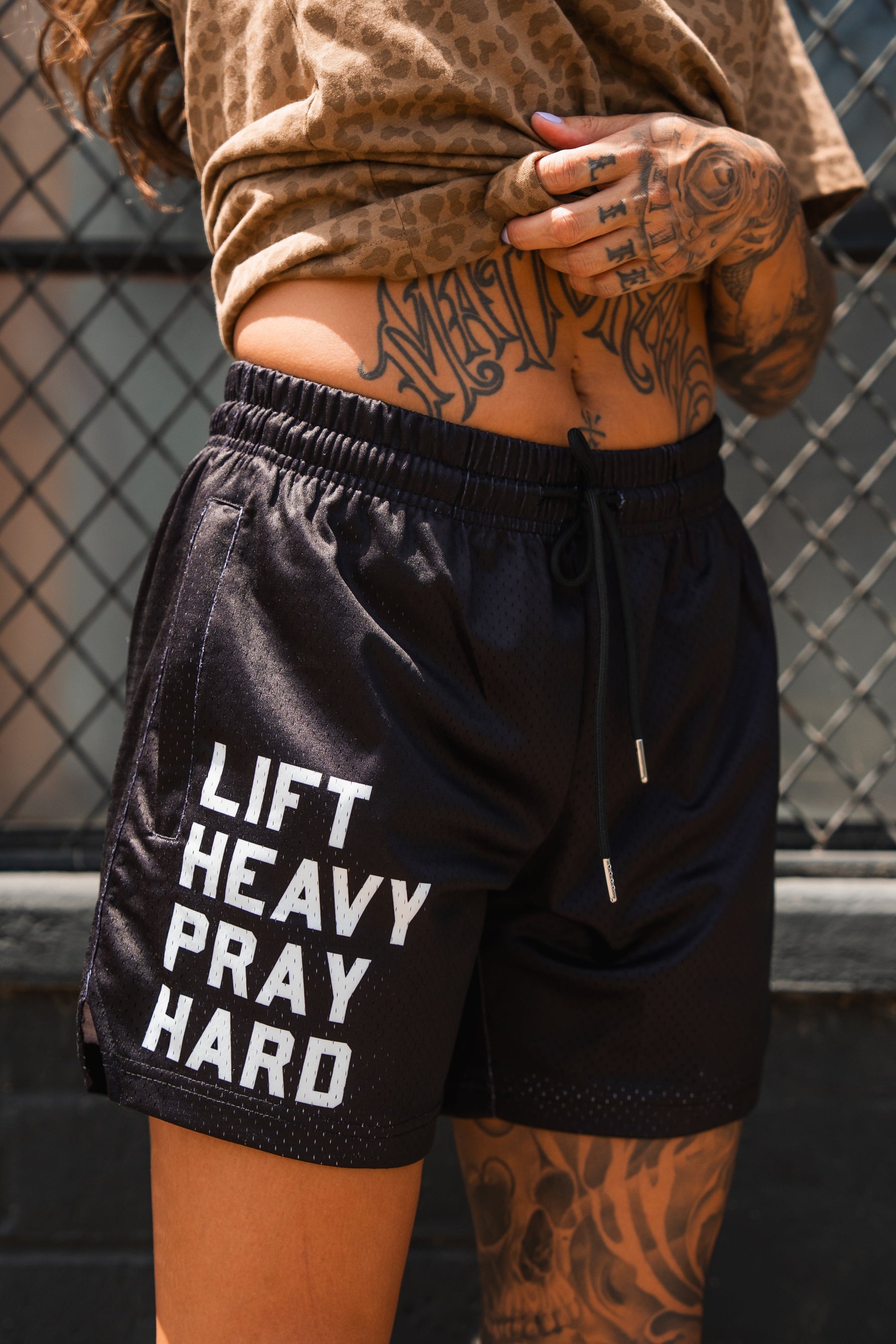 A person with tattooed arms wears Tango Charlie Apparel's Men's Mesh Lifestyle Shorts | 5.5" - Lift Heavy Pray Hard Black, featuring white "LIFT HEAVY PRAY HARD" text, while standing in front of a metal fence.