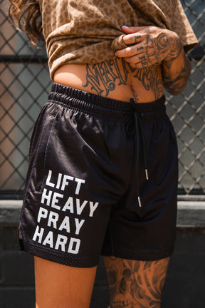A person with tattooed arms wears Tango Charlie Apparel's Men's Mesh Lifestyle Shorts | 5.5" - Lift Heavy Pray Hard Black, featuring white "LIFT HEAVY PRAY HARD" text, while standing in front of a metal fence.