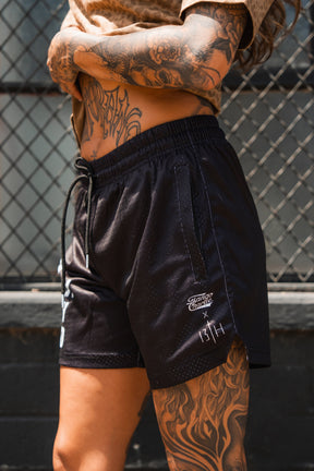 A person with tattooed arms, hands, and torso wears Tango Charlie Apparel Men's Mesh Lifestyle Shorts | 5.5"- Lift Heavy Pray Hard Black and a brown shirt outdoors in front of a chain-link fence.