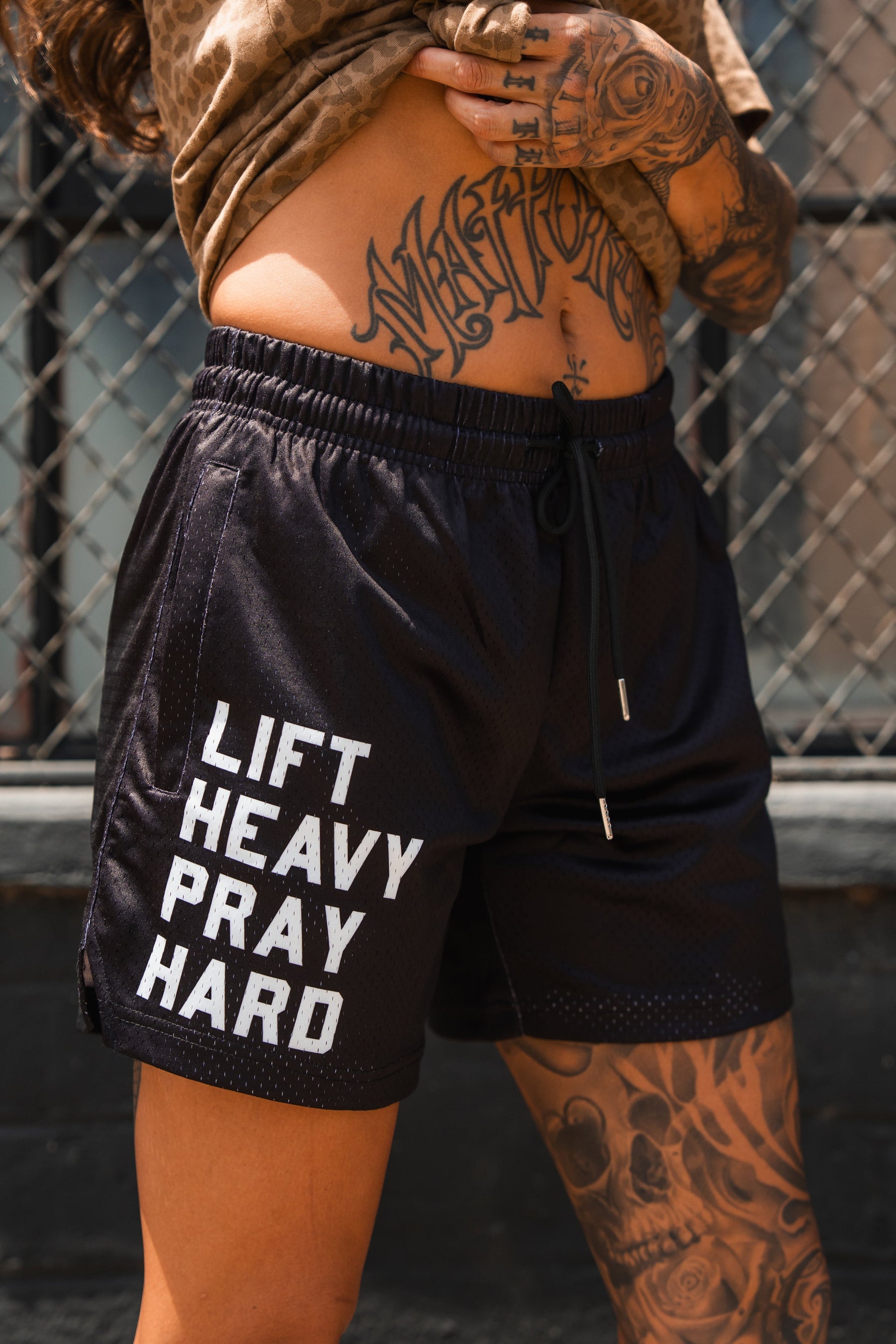 A tattooed person stands by a chain-link fence wearing Tango Charlie Apparel's Men's Mesh Lifestyle Shorts | 5.5"- Lift Heavy Pray Hard Black, featuring "LIFT HEAVY PRAY HARD" in white letters.