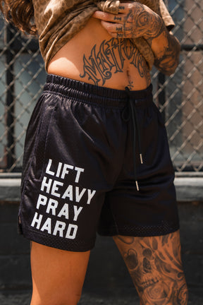 A tattooed person stands by a chain-link fence wearing Tango Charlie Apparel's Men's Mesh Lifestyle Shorts | 5.5"- Lift Heavy Pray Hard Black, featuring "LIFT HEAVY PRAY HARD" in white letters.