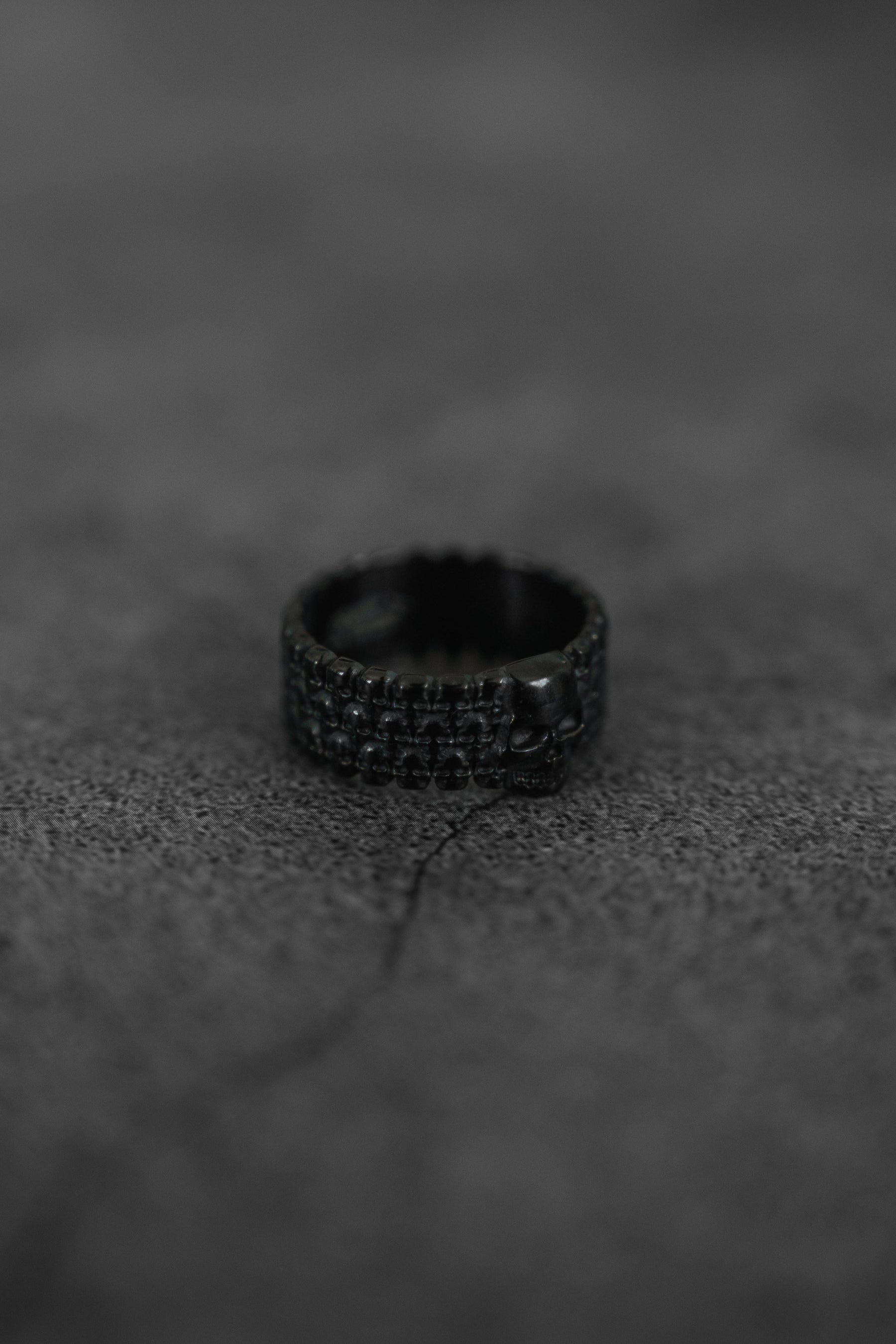 The Kill Switch Blackout Ring by Relentless Betrayal, featuring a small skull detail and matte black finish, sits on a rugged dark surface, evoking a bold and dramatic vibe.