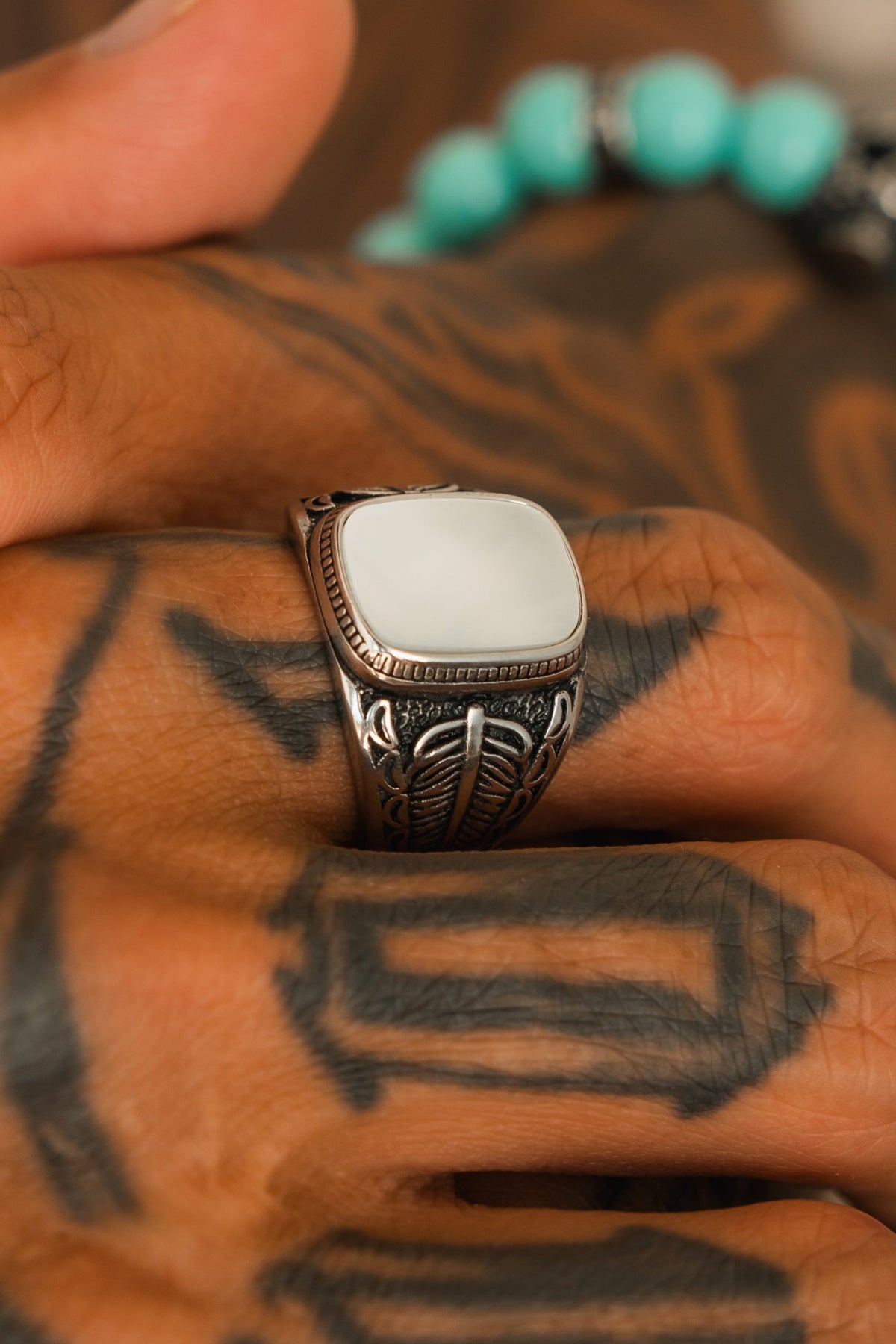 A hand with black geometric tattoos wears the Relentless Betrayal Desire Opal Ring, a handcrafted silver piece with an ornate design and rectangular white opal. Turquoise beads are softly blurred in the background.