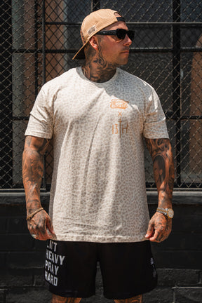 A tattooed man in sunglasses, a beige animal-print tee, tan cap, and Tango Charlie Apparel Men's Mesh Lifestyle Shorts (5.5" - Lift Heavy Pray Hard Black) stands before a black metal fence.