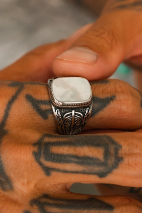 A hand with black geometric tattoos wears the Relentless Betrayal Desire Opal Ring, featuring a large, smooth white stone. The visible thumb and fingers highlight the handcrafted jewelry’s intricate details.