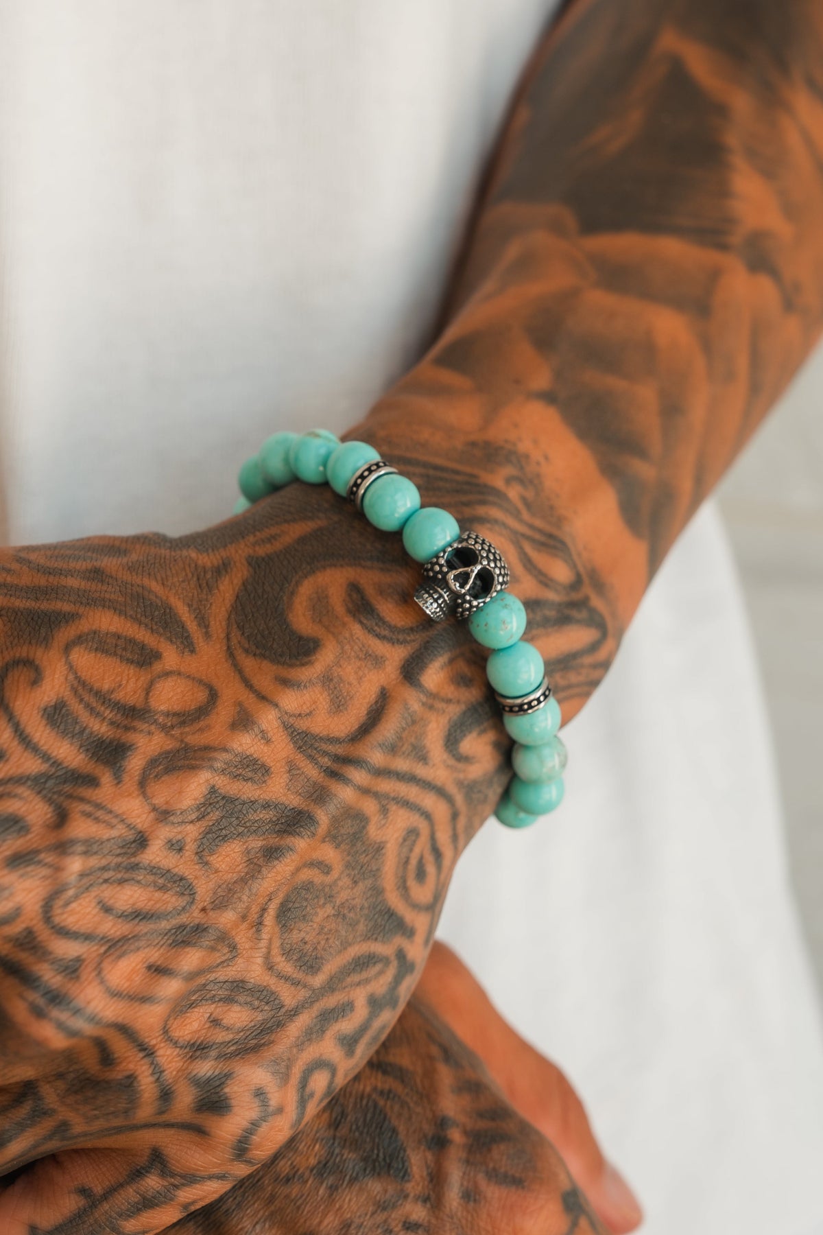 A close-up of tattooed arms in a white shirt, hands crossed, wearing the Relentless Betrayal Day of the Dead Bead Bracelet—turquoise beads with a decorative silver skull charm.