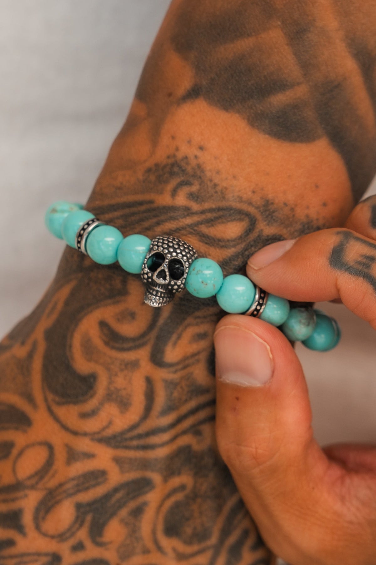 A tattooed hand holds the Relentless Betrayal Day of the Dead Bead Bracelet, crafted with turquoise beads, silver skull charm, and decorative spacers, set against a brown tattooed background.