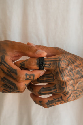 Tattooed hands place a Relentless Betrayal Kill Switch Blackout Ring on the ring finger, showcased against a light fabric background.