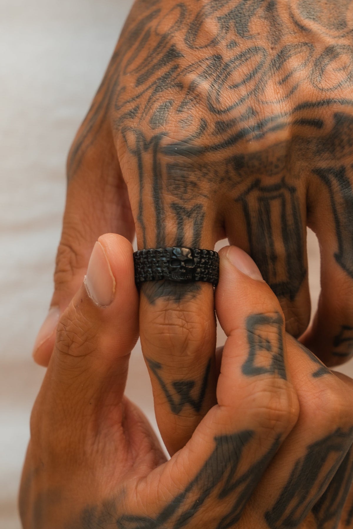 Tattooed hands, one holding the other’s finger, display the Relentless Betrayal Kill Switch Blackout Ring—a matte black piece with a skull design—its bold details stand out against a softly blurred background.