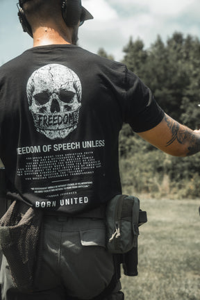 A person stands outdoors in a grassy area with trees, wearing tactical gear and the Born United Private Label "Freedom of Speech" t-shirt—featuring a bold skull graphic and 'FREEDOM OF SPEECH UNLESS' text—making a striking statement about free expression.