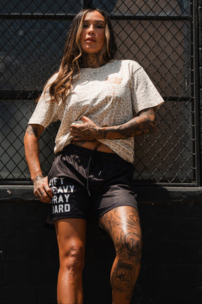 A tattooed woman stands confidently by a black metal fence, wearing a beige patterned tee and Tango Charlie Apparel’s Men's Mesh Lifestyle Shorts | 5.5"–Lift Heavy Pray Hard Black, with sunlight illuminating her face and tattoos.