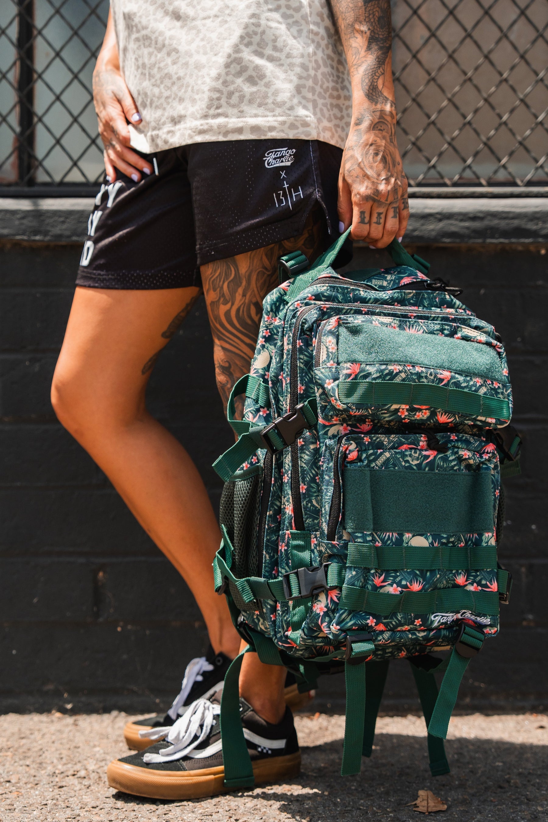 A tattooed individual stands on a sidewalk holding the Aloha AR - Tac Pac 25L by Tango Charlie Apparel, a green, floral-patterned waterproof backpack. They're dressed in black shorts, a patterned shirt, and black sneakers; a wire fence and brick wall are behind them.