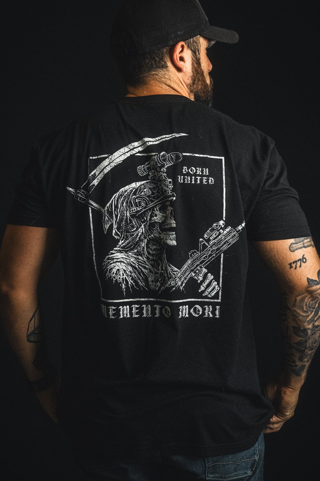 A man facing away wears a black cap and a black T-shirt from Born United Private Label’s “Memento Mori” collection, featuring a striking graphic of a skeleton warrior and the text “Memento Mori” and “Born United” on the back.