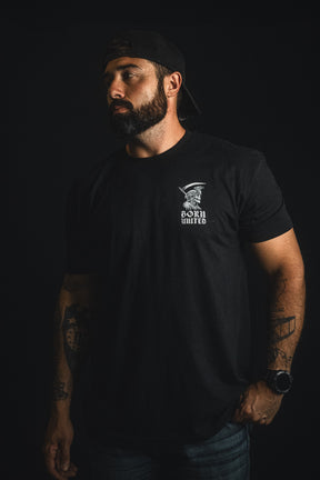 A bearded, tattooed man in a black Born United Private Label "Memento Mori" T-shirt and backwards cap stands against a dark background, glancing to the side—his thoughtful expression echoing the collection’s reminder of life’s impermanence.