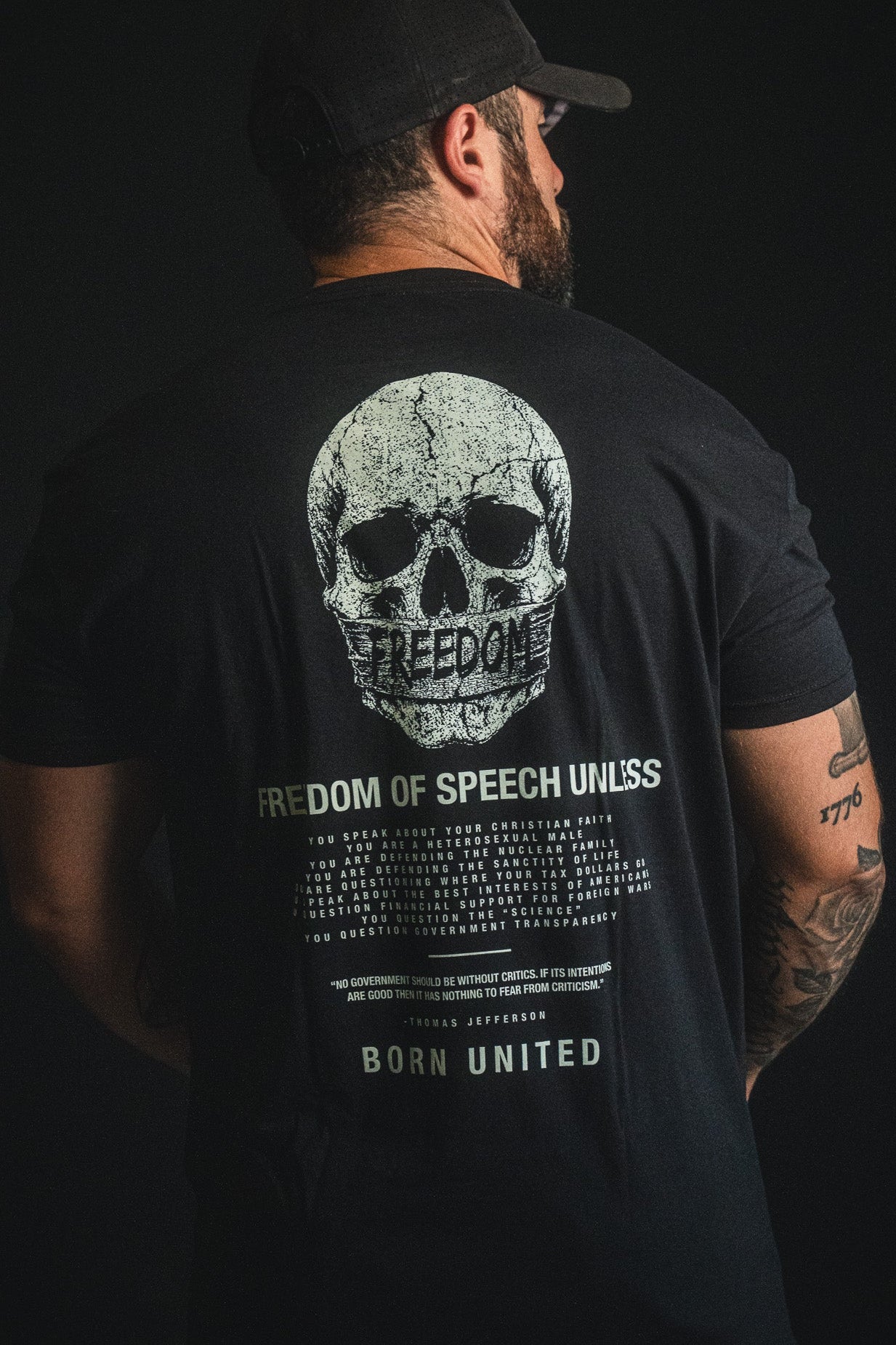 A man stands against a dark background, showcasing his right arm tattoos while wearing the Born United Private Label "Freedom of Speech" T-shirt—a bold statement of personal beliefs and free expression.