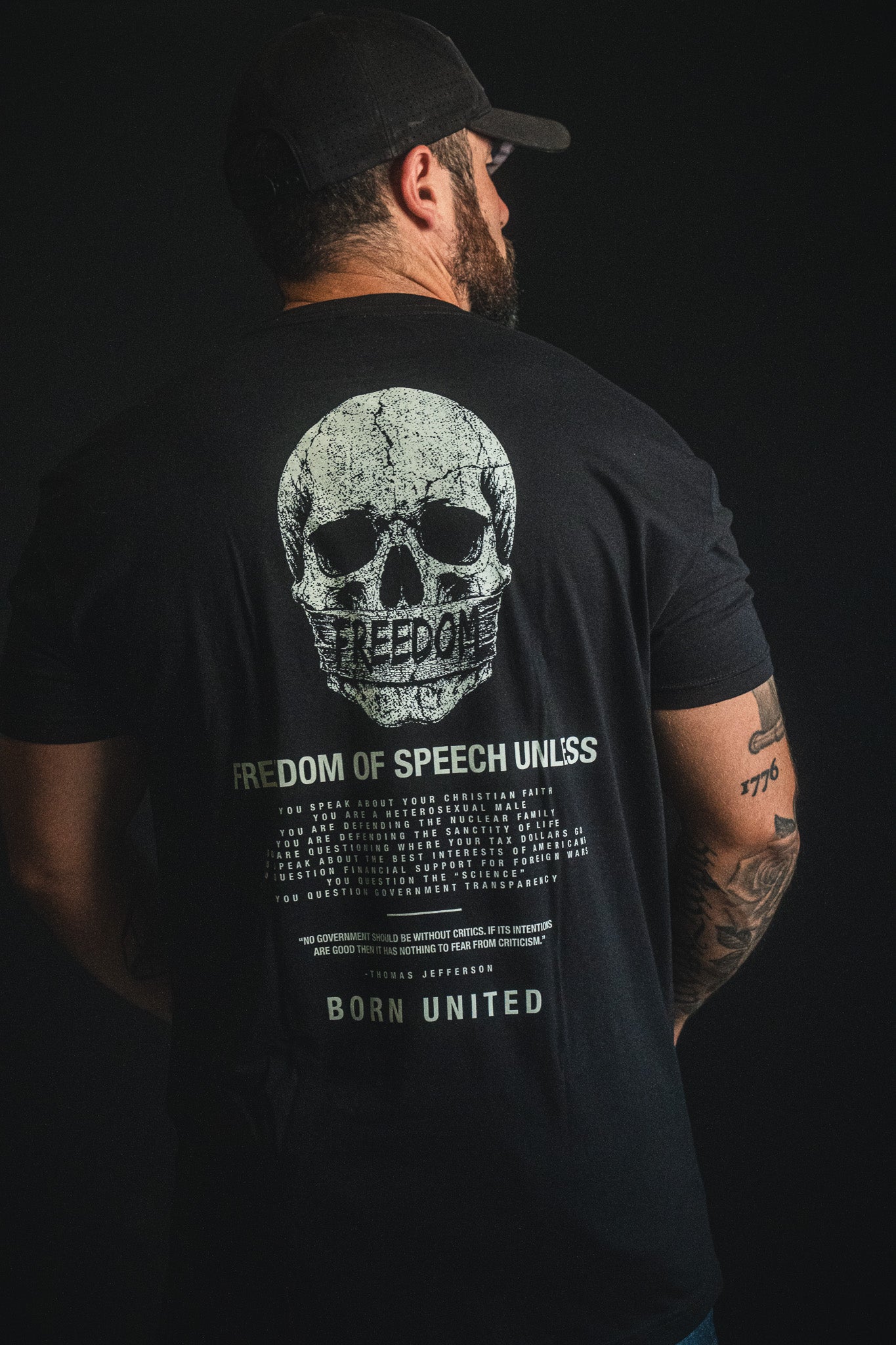A man stands against a dark background, showcasing his right arm tattoos while wearing the Born United Private Label "Freedom of Speech" T-shirt—a bold statement of personal beliefs and free expression.