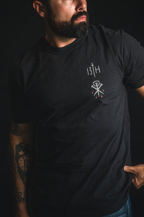 A bearded man wearing the 13th Nothing But the Blood Premium Tee—a black Christian t-shirt featuring "13TH" and small tattoo-style graphics on the chest—stands against a dark background, showcasing a unique blend of modern style and spiritual inspiration.