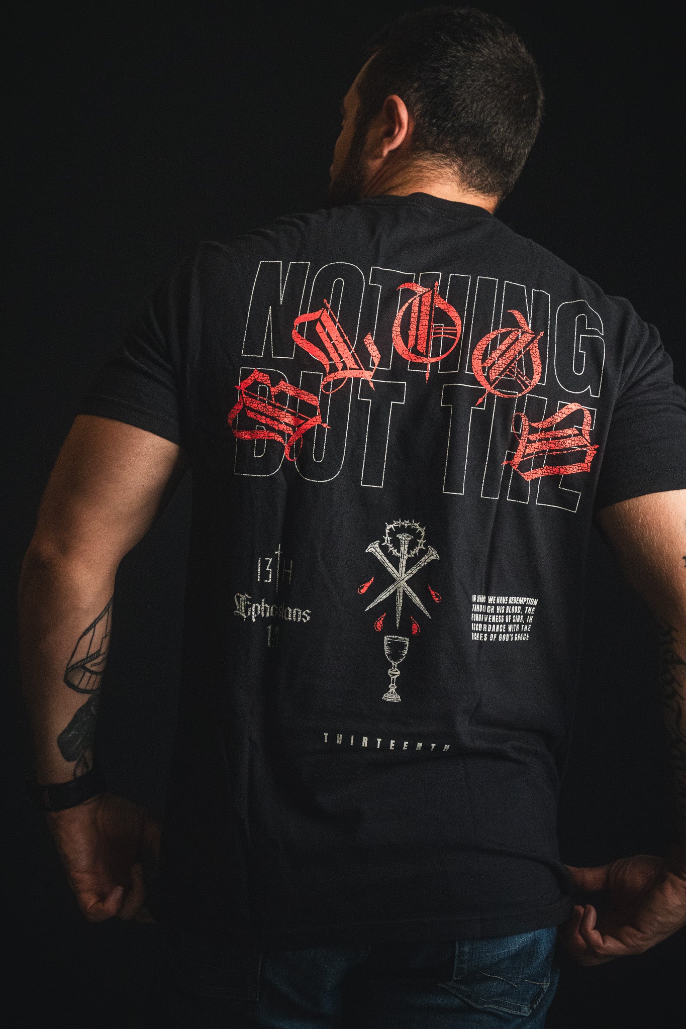 A man stands with his back to the camera, wearing the 13th brand’s Nothing But the Blood Premium Tee, a black shirt featuring bold red and white graphic text and symbols.
