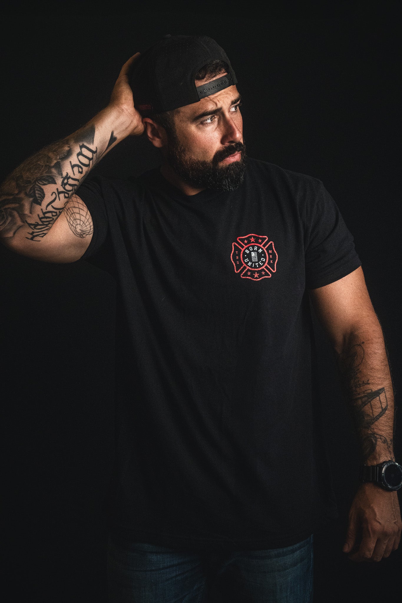 A man stands against a dark background, wearing a black t-shirt featuring The Fireman's Prayer by Born United Private Label and a backward cap, his tattooed arm resting behind his head. This powerful image makes an ideal firefighter gift or inspiration for a heartfelt firefighter poem.
