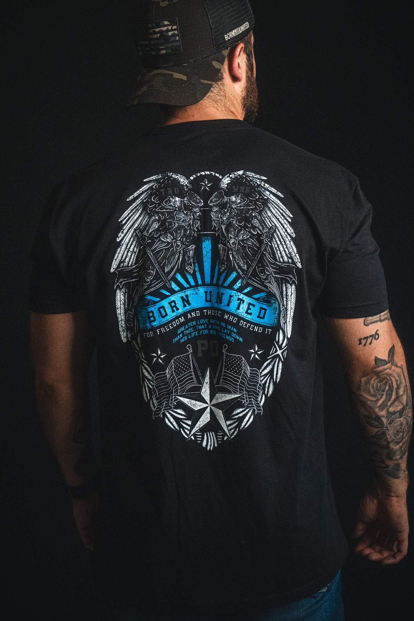 A man is wearing a black Born United Private Label "Back the Badge" cap and a matching black T-shirt featuring an eagle, wings, stars, and the phrases "Born United" and "For freedom and those who defend it" on the back. A tattoo is visible on his left arm.
