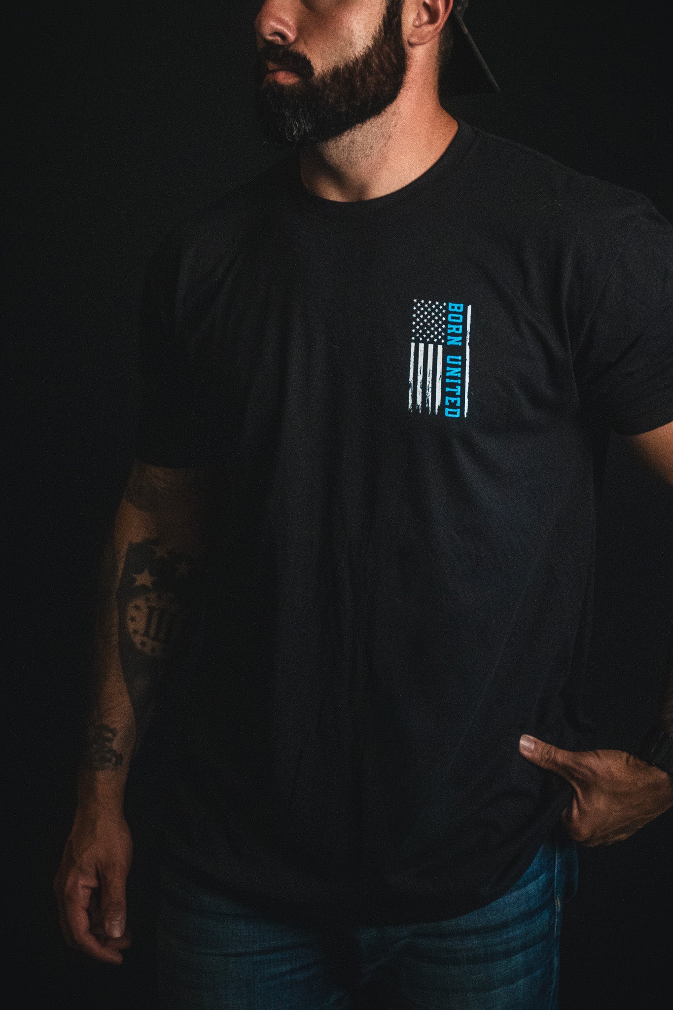 A man wearing the Born United Private Label "Back the Badge" black t-shirt featuring a blue line American flag and "Born United" text stands against a dark background.