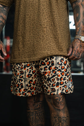 A tattooed individual wears Tango Charlie Apparel Men's Mesh Lifestyle Shorts | 5.5"- Loaded Leopard with a brown leopard print shirt and a gold watch, standing against a dark, blurred background.