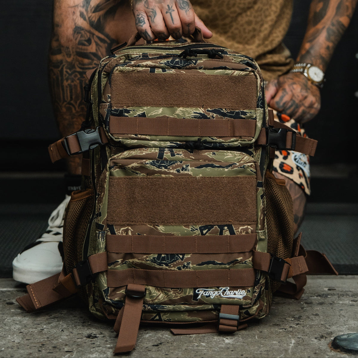 A tattooed individual sits outdoors on concrete, holding the Tango Charlie Apparel Snipes & Stripes - Tac Pac 25L backpack featuring brown straps and mesh pockets.