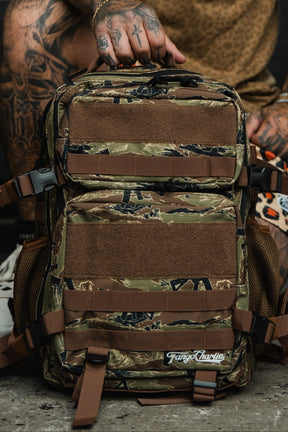A person with tattooed hands holds the Snipes & Stripes - Tac Pac 25L, a camouflage backpack with brown straps and mesh pockets. The Tango Charlie Apparel logo is visible on the durable pack, which sits on a concrete floor.