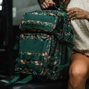 A person with tattooed hands sits on a bench, holding the Tango Charlie Apparel Aloha AR - Tac Pac 25L, a large green backpack with a floral and leaf pattern. They wear a light-colored shirt and shorts.