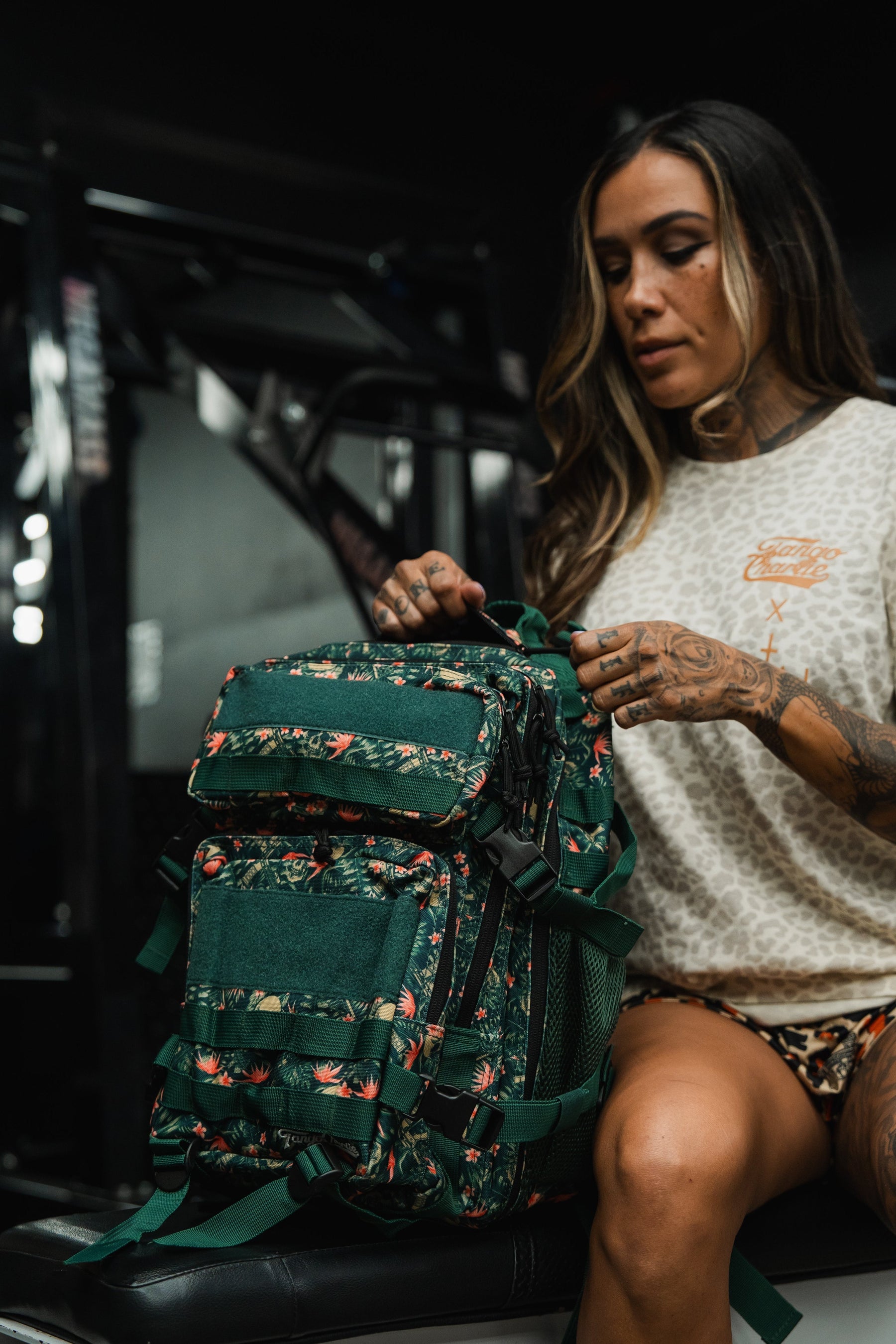 A woman with long hair and tattoos sits indoors, holding the Tango Charlie Apparel Aloha AR - Tac Pac 25L backpack with a green floral pattern on her lap. She wears a beige shirt and shorts in a dimly lit gym setting.