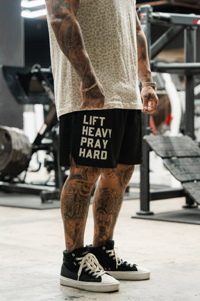 A tattooed person stands in a gym, wearing a leopard print shirt, gold bracelets, black high-top sneakers, and Tango Charlie Apparel Men's Mesh Lifestyle Shorts | 5.5"- Lift Heavy Pray Hard Black. Gym equipment is seen in the background.