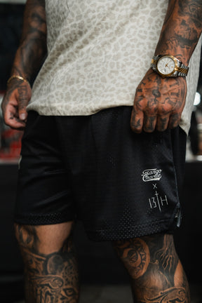 A tattooed person wears a leopard print shirt, gold watch, and black Men's Mesh Lifestyle Shorts 5.5" – Lift Heavy Pray Hard by Tango Charlie Apparel, standing indoors.
