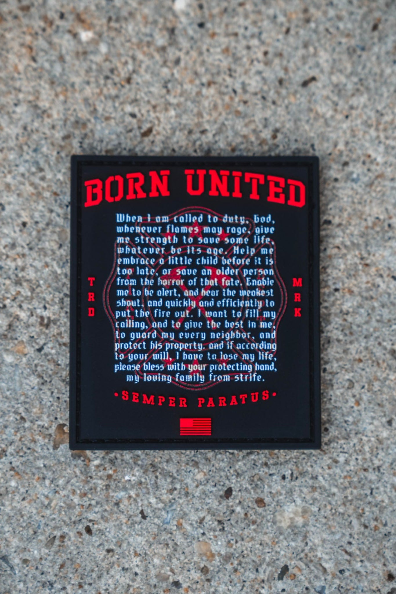 A Fireman's Prayer PVC Patch from Born United Private Label, featuring a black and red 3D design, rests on a gravel surface; it showcases the words "Born United," a prayer about duty and service, the motto "Semper Paratus," and an American flag graphic at the bottom.