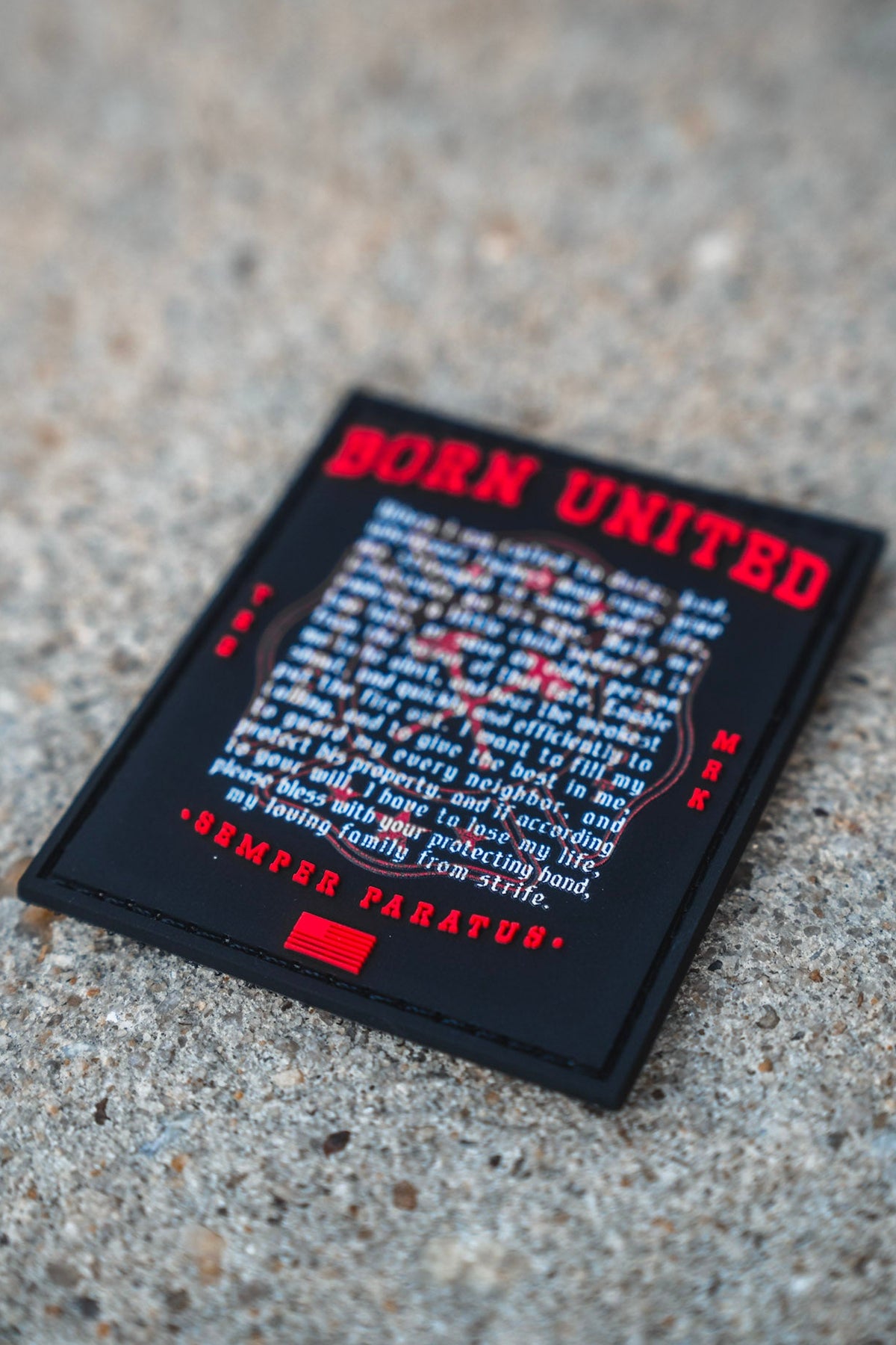 A rectangular 3 inch Fireman's Prayer PVC Patch by Born United Private Label, featuring red and white text, rests on a rough concrete surface. The patch displays a motivational message with the phrase SEMPER PARATUS at the bottom.