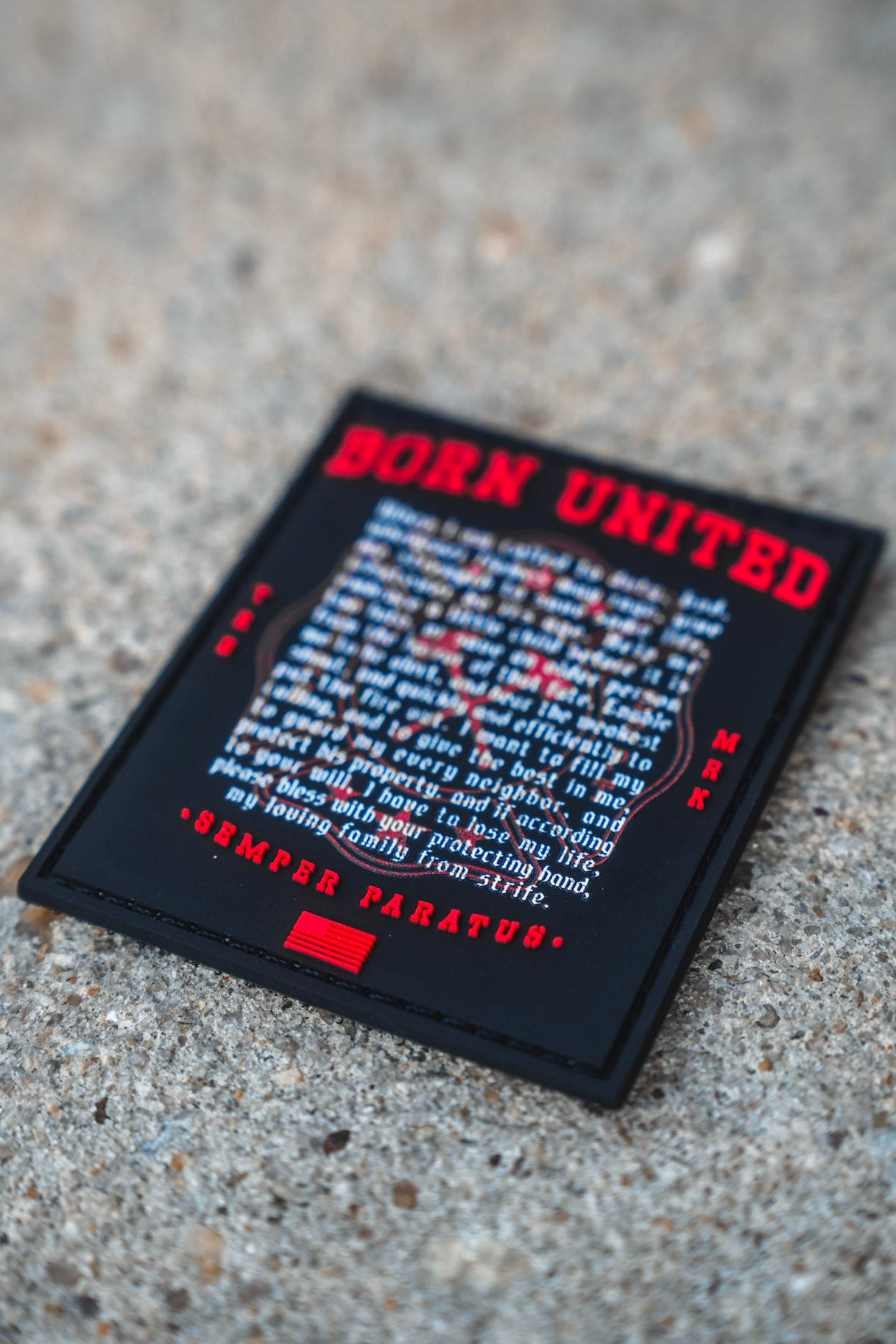 A rectangular 3 inch Fireman's Prayer PVC Patch by Born United Private Label, featuring red and white text, rests on a rough concrete surface. The patch displays a motivational message with the phrase SEMPER PARATUS at the bottom.