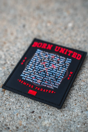 A rectangular 3 inch Fireman's Prayer PVC Patch by Born United Private Label, featuring red and white text, rests on a rough concrete surface. The patch displays a motivational message with the phrase SEMPER PARATUS at the bottom.