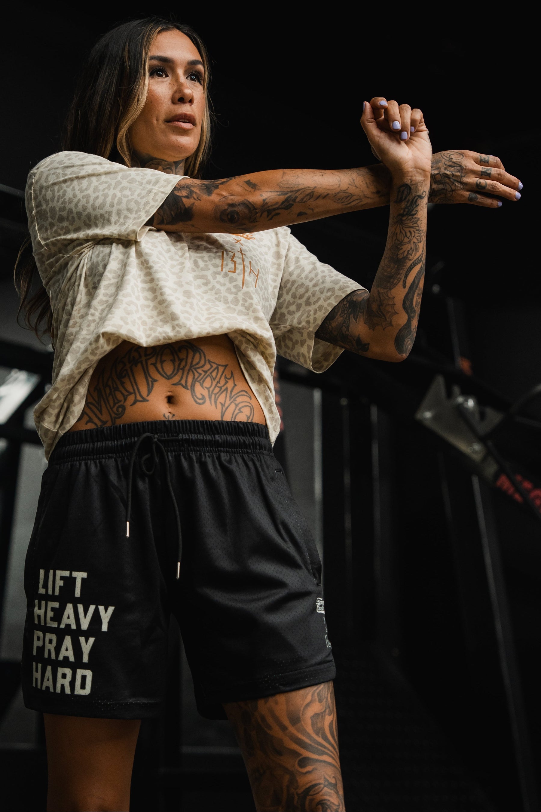 A tattooed woman wearing Tango Charlie Apparel's Men's Mesh Lifestyle Shorts 5.5" in Lift Heavy Pray Hard Black and a light-patterned top stretches her arm across her chest in a vibrant gym setting.