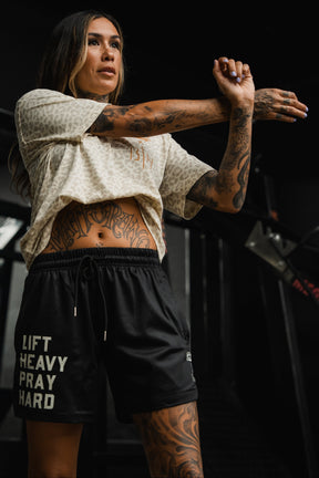 A tattooed woman wearing Tango Charlie Apparel's Men's Mesh Lifestyle Shorts 5.5" in Lift Heavy Pray Hard Black and a light-patterned top stretches her arm across her chest in a vibrant gym setting.