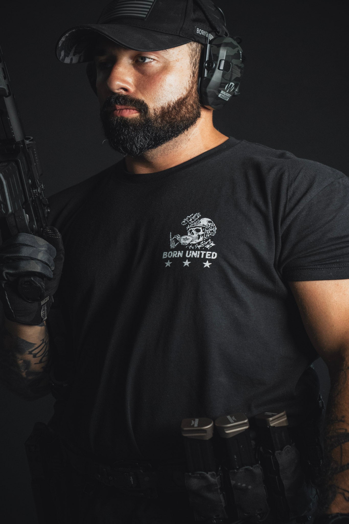 A bearded man stands against a dark background, wearing a black t-shirt, tactical gloves, and headphones while holding a firearm—looking more prepared for action than for enjoying the Tea Party by Born United Private Label.