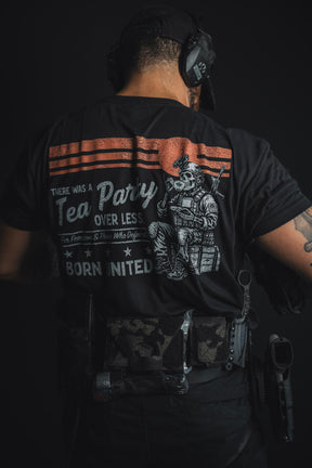 A man dressed in tactical gear and headphones stands with his back to the camera, showcasing the ‘Tea Party’ T-shirt from Born United Private Label, which features the text: “There was a Tea Party over less. Born united.” — a modern nod to historic rebellion.