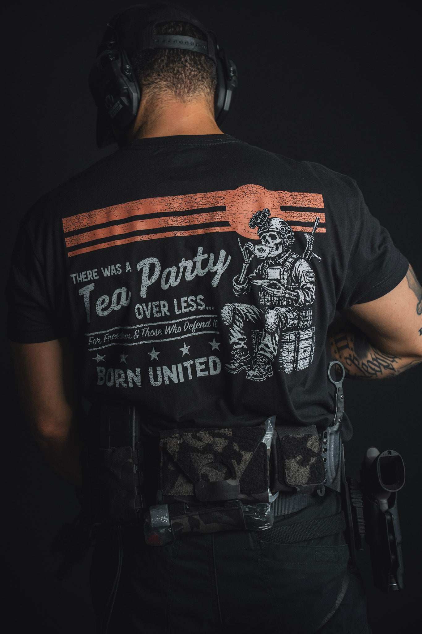 A person in tactical gear faces away, wearing the Born United Private Label “Tea Party” shirt featuring a graphic of a soldier drinking tea from a tea set and the text, There was a Tea Party over less… Born United.