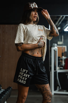 A tattooed woman poses in a gym wearing Tango Charlie Apparel’s Men's Mesh Lifestyle Shorts | 5.5"- Lift Heavy Pray Hard Black, paired with a leopard-print shirt and brown cap, holding the brim as she lifts her shirt; fitness equipment fills the background.