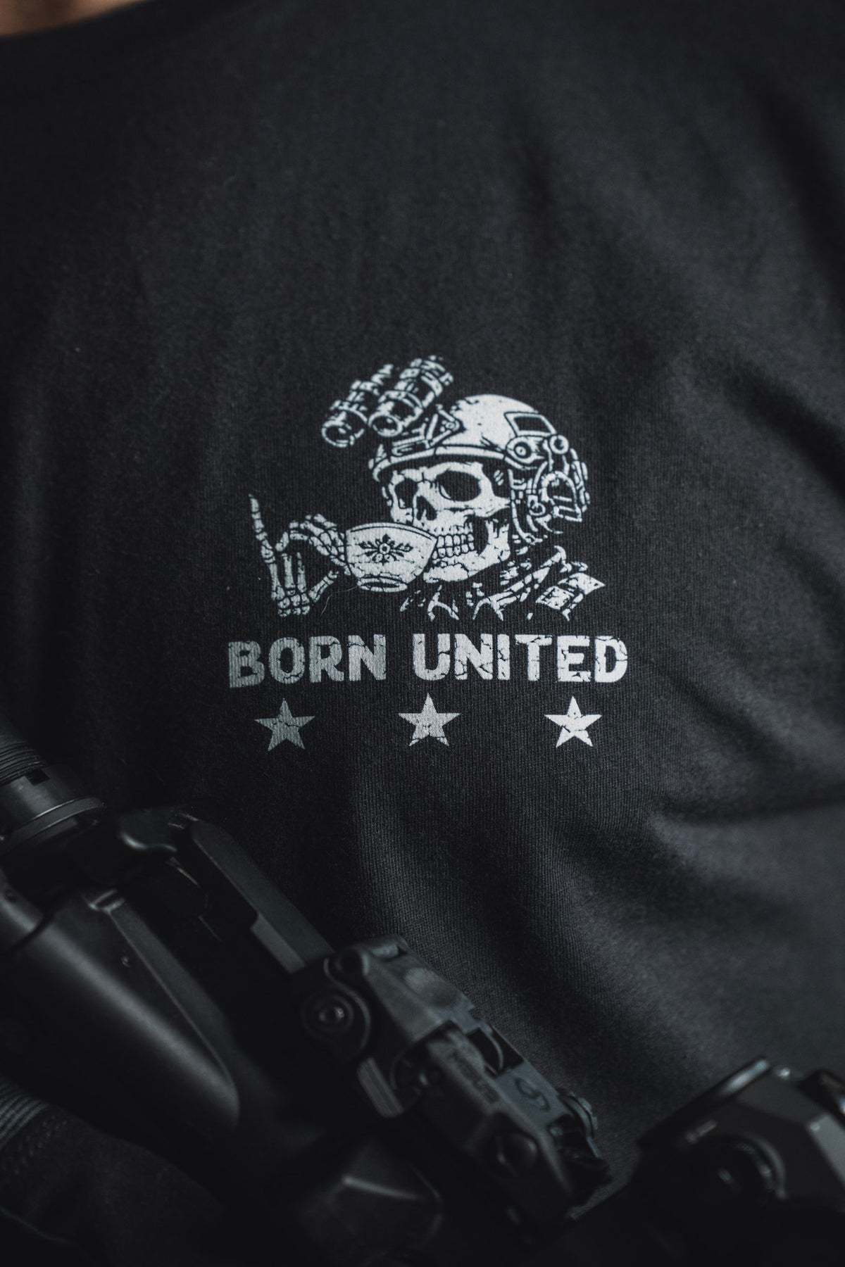 Introducing the “Tea Party” tee from Born United Private Label: a bold black shirt featuring a graphic of a skull in tactical gear flashing a hand signal above the words BORN UNITED and three stars. With part of a firearm visible in the foreground, this striking design is perfect for those who like their party supplies with an edge.
