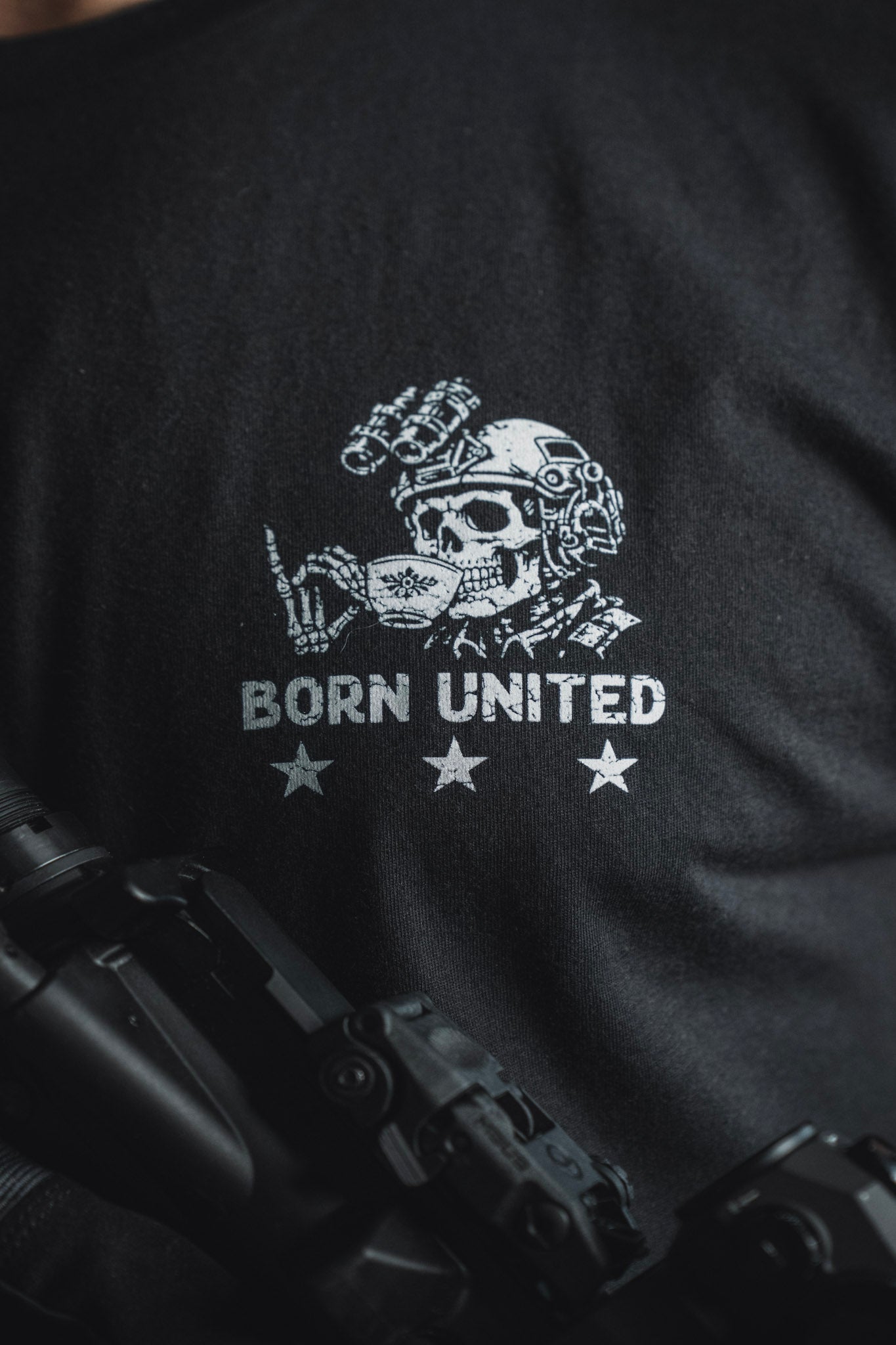 Introducing the “Tea Party” tee from Born United Private Label: a bold black shirt featuring a graphic of a skull in tactical gear flashing a hand signal above the words BORN UNITED and three stars. With part of a firearm visible in the foreground, this striking design is perfect for those who like their party supplies with an edge.