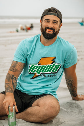 A bearded, tattooed man smiles on the beach with a can in hand, wearing Tango Charlie Apparel’s “Tequila Is My Gatorade - Berry Blue” shirt. People and the ocean blur behind him, capturing the perfect summer vibe.