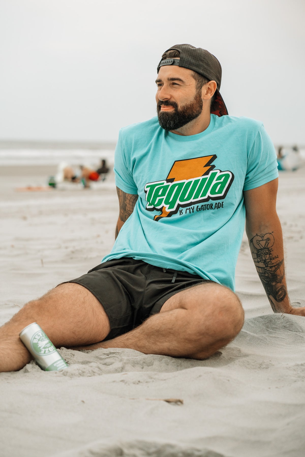 A bearded man in a Tango Charlie Apparel Tequila Is My Gatorade - Berry Blue shirt and black shorts relaxes on the beach, a drink can by his side as he enjoys bold colors, summer vibes, and an ocean view.