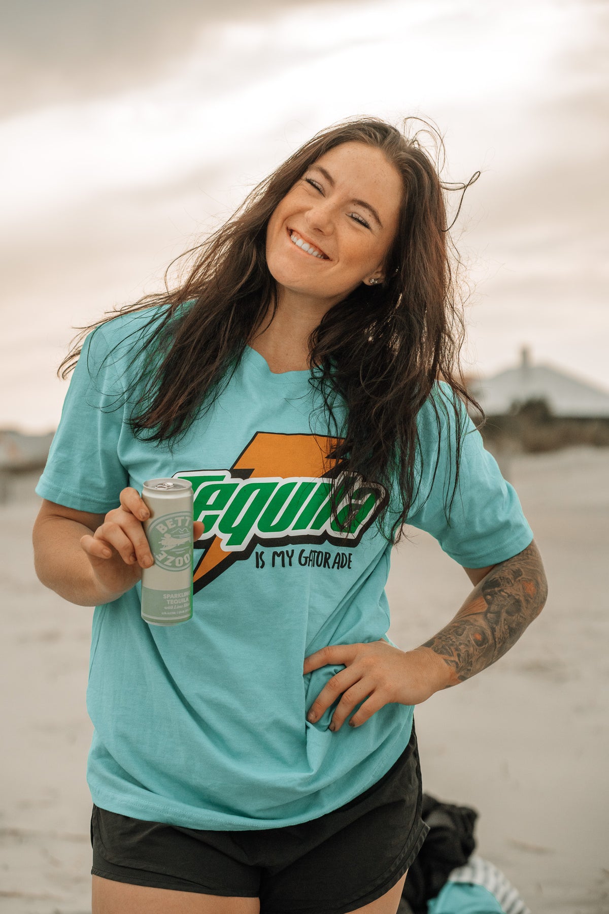 A smiling woman stands on the beach holding a canned drink, wearing black shorts and a light blue Tango Charlie Apparel tee reading Tequila Is My Gatorade - Berry Blue—perfect for summer vibes, with her left forearm tattoo visible.